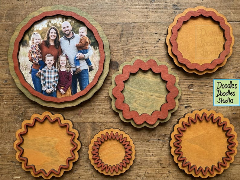 Peut inclure: Une collection de cadres circulaires en bois aux bords festonn&eacute;s dans des tons marron, beige et vert. Un cadre contient une photographie d'une famille de cinq personnes. Un petit panneau carr&eacute; bleu avec le texte Doodles Doodles Studio est visible.