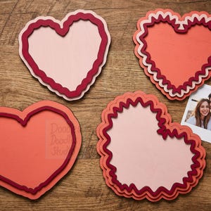 Peut inclure: Quatre d&eacute;coupes en bois en forme de c&oelig;ur superpos&eacute;es dans des tons de rose, corail et rouge fonc&eacute;. Les c&oelig;urs pr&eacute;sentent des bords festonn&eacute;s ou lisses et des motifs de bordure vari&eacute;s. Une petite photo carr&eacute;e d'un couple est plac&eacute;e pr&egrave;s du c&oelig;ur.