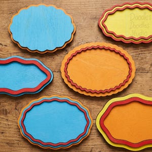 Peut inclure: Six plaques en bois aux bords festonn&eacute;s de couleur bleue, jaune et orange. Les plaques pr&eacute;sentent des motifs superpos&eacute;s avec des bordures rouges ou jaunes. Une plaque jaune porte le texte Doodles Doodles Studio et un logo affiche ce texte.