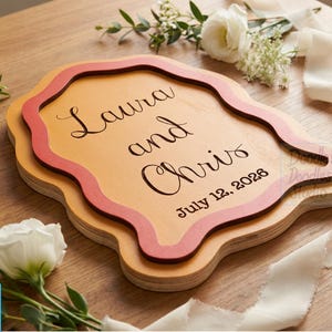 Peut inclure: Une plaque en bois superpos&eacute;e de forme organique ondul&eacute;e, compos&eacute;e d'une base en bois clair et d'une couche sup&eacute;rieure de couleur corail fonc&eacute;. Le texte Laura and Chris et la date July 12, 2026 sont grav&eacute;s en noir dans une police cursive &eacute;l&eacute;gante.