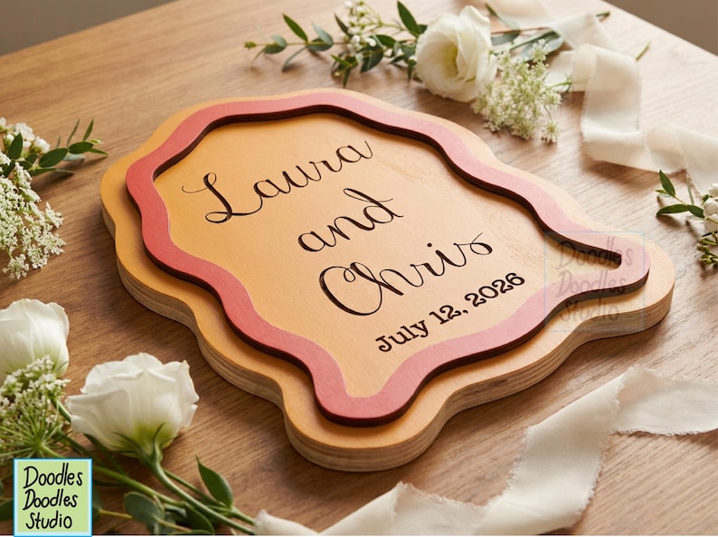 Peut inclure: Une plaque en bois superpos&eacute;e de forme organique ondul&eacute;e, compos&eacute;e d'une base en bois clair et d'une couche sup&eacute;rieure de couleur corail fonc&eacute;. Le texte Laura and Chris et la date July 12, 2026 sont grav&eacute;s en noir dans une police cursive &eacute;l&eacute;gante.