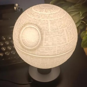May include: A white ceramic lamp shaped like the Death Star from Star Wars. The lamp has a textured surface and a black base.
