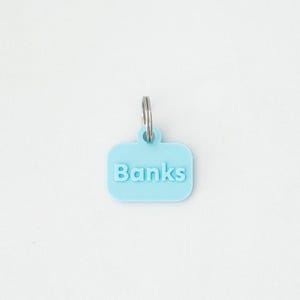 May include: A light blue pet tag with the name "Banks" in white, attached to a silver metal ring. The tag is rectangular with rounded corners and is set against a white background.