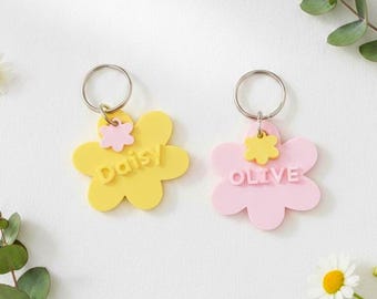 May include: Two flower-shaped keychains, one yellow with "Daisy" and the other pink with "Olive" in white text. Each has a smaller flower accent. The keychains are on a white surface with greenery.