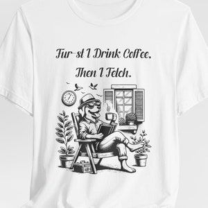 Może przedstawiać: Biały T-shirt z czarno-białą ilustracją psa siedzącego na krześle, pijącego kawę i czytającego książkę. Na koszulce nadrukowany jest tekst "First I Drink Coffee, Then I Fetch."