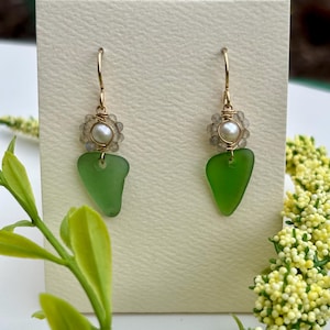 Puede incluir: Pendientes hechos a mano con un colgante de vidrio verde mar en forma de lágrima. Cada pendiente tiene una pequeña perla y un grupo de cuentas transparentes, suspendidas de un gancho dorado.
