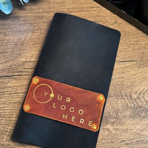 May include: Black leather notebook with a rectangular, brown leather patch. The patch is embossed with the words "YOUR LOGO HERE" in gold lettering. The notebook is on a wooden surface.
