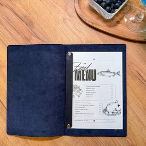 Könnte beinhalten: Offener, marineblauer Leder-Menühalter mit einem gedruckten Menü im Inneren. Das Menü zeigt die Worte "Food MENU" oben, mit Illustrationen von Fisch, Gemüse und einem Brathähnchen. Der Halter befindet sich auf einer Holzoberfläche.