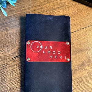 May include: A rectangular dark navy blue notebook featuring a horizontal red leather patch centered on the front cover. The red patch is secured with four silver-toned metal rivets and displays the white text YOUR LOGO HERE with a circular icon.