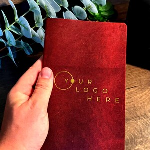 Op de afbeelding: Een dieprood, rechthoekig notitieboek met een matte, getextureerde kaft. De voorzijde is voorzien van een goudkleurige cirkelvormige afbeelding en de tekst "YOUR LOGO HERE" in een strak, schreefloos lettertype.