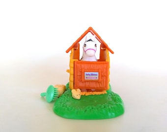 Könnte beinhalten: Ein Spielzeugstall aus Kunststoff mit orangefarbenem Dach und gelben Wänden, in dem eine weiße Pferdefigur mit rosa Zaumzeug steht. Eine kleine beige Mausfigur und eine grüne Kunststoffbürste liegen auf der grünen Basis. Das Schild zeigt Baby Mouse.