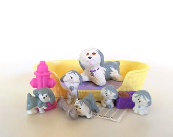 May include: A collection of miniature toy dogs in a yellow bed, with a pink fire hydrant and a bone. The dogs are white and gray, with a purple dog bed and a purple food bowl. The toys are from the 1992 Kenner collection.