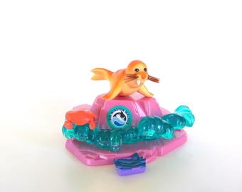 May include: An orange walrus figurine with brown whiskers and white tusks perched on a pink rock base. The base features a circular sticker with an orca graphic and the text SIR WINSTON WALRUS, a small orange crab, translucent blue waves, and a purple crate.