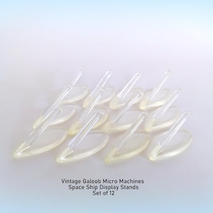 May include: A set of 12 clear plastic display stands for toy spaceships. Each stand features an oval base with an angled support arm. The text below reads: Vintage Galoob Micro Machines Space Ship Display Stands Set of 12.