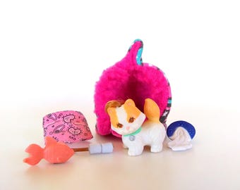 May include: A small white and orange plastic cat figurine with a green collar, positioned in front of a bright pink fuzzy cave-like structure. Accessories include a pink patterned pillow, a small orange fish toy, and a blue and white net.