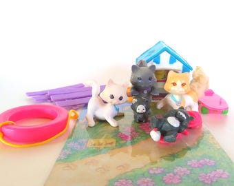 May include: A collection of miniature plastic cat figurines including a white cat, a grey cat, an orange and white cat, and two smaller black kittens. Accessories include a blue and white striped house, a pink life preserver, and a purple fence.