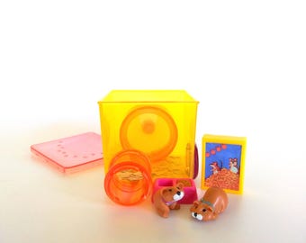 Pode incluir: Uma gaiola de hamster de plástico amarelo translúcido com um túnel laranja, uma tigela de comida rosa, uma tampa retangular rosa e um painel gráfico com moldura amarela mostrando dois hamsters. Duas pequenas figuras de hamster marrom estão à frente.