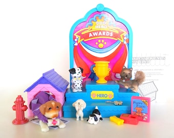 Könnte beinhalten: Ein farbenfrohes Spielset mit Miniatur-Hundefiguren und Zubehör. Das Set enthält ein Hundehäuschen, einen Hydranten, eine Trophäe und ein Schild mit der Aufschrift "Dog Hero Awards". Die Hunde sind in verschiedenen Posen, für fantasievolles Spielen.