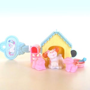 May include: A miniature yellow doghouse with a light blue roof, a pink mailbox, and three small plastic dog figurines. One dog is inside the house, while two others sit in front. A light blue key-shaped accessory features a graphic of a white dog.