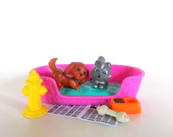 May include: A pink plastic pet bed containing a brown puppy and a gray kitten figurine. Beside the bed is a yellow fire hydrant, an orange food bowl, a white bone toy, and a small rectangular paper sheet with 1992 KENNER text printed.
