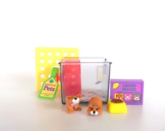 Könnte beinhalten: Ein Miniatur-Haustier-Spielset mit einem transparenten Kunststoffkäfig, einem roten Laufrad, zwei kleinen braunen Tierfiguren, einem gelben Futternapf, einer lila Schachtel mit der Aufschrift Critter Snacks und einem grünen Anhänger.