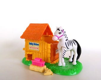 May include: A toy set featuring a white and black zebra figurine with a pink bridle, standing on a green base. The set includes a small orange and yellow barn with the text "Baby Zebra" and a pink brush.