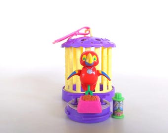 Könnte beinhalten: Ein farbenfrohes Spielzeug, das einen Vogel in einem lila und gelben Käfig darstellt. Der Vogel ist rot mit blauen und gelben Akzenten. Eine rosa Futterschale und ein grüner Behälter mit einem Vogelmuster sind ebenfalls vorhanden. Der Käfig hat eine lila Oberseite und einen rosa Griff.