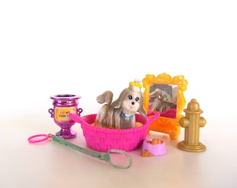 May include: A toy set featuring a small Shih Tzu dog figure with a yellow bow sitting in a pink basket. Accessories include a purple trophy with the text I LOVE MY SHIH TZU, a yellow mirror, a gold fire hydrant, a pink food bowl, and a green wand.