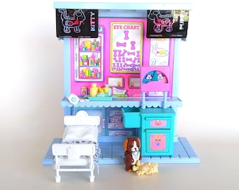 May include: A colorful toy pet clinic with a bed, eye chart, and various accessories. The clinic features a blue and pink color scheme, with a brown and white dog figurine and a yellow dog figurine. The sign says "KITTY" and "PUPPY".