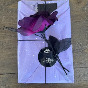 May include: A wrapped gift in lavender paper with silver flecks, tied with black twine. A large, dark purple rose and black leaves adorn the package. A black circular sticker with the text "in my VAMPIRE ERA" is visible.