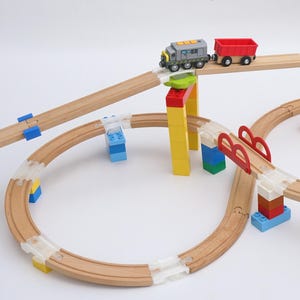 Op de afbeelding: Een houten speelgoedtreinset met een grijze en rode trein en een rode goederenwagon. De baan bevat kleurrijke steunen en een brug. Tekst op de afbeelding zegt "Ein Adapter - für Aufstieg, Abstieg, Anfang & Ende einer Rampe - maximale Flexibilität."