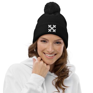 Op de afbeelding: Zwarte beanie met een wit geborduurd kruisontwerp. De beanie heeft een geribbelde textuur en een zwarte pompon. De persoon draagt een witte hoodie. Een winteraccessoire.