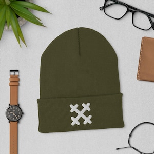 Op de afbeelding: Olijfgroene beanie met een wit geborduurd kruisontwerp op de voorkant. De hoed is bij de rand gevouwen. Een horloge, een bril en een portemonnee zijn ook zichtbaar. De hoed ligt op een lichtgrijze ondergrond.