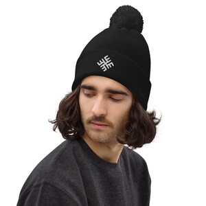 Op de afbeelding: Zwarte beanie met een wit geborduurd ontwerp en een zwarte pompon. De beanie is gemaakt van geribbelde gebreide stof. De persoon op de foto draagt een donkergrijs crewneck shirt.