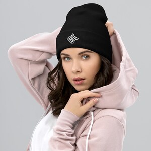 Op de afbeelding: Zwarte beanie met een wit geborduurd geometrisch ontwerp. De beanie is gemaakt van zacht, geribbeld breiwerk. Een stijlvol winteraccessoire voor koud weer.