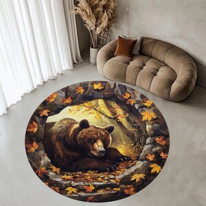 May include: Circular rug with a detailed illustration of a brown bear in a forest setting with autumn leaves. The rug depicts a bear in a cave, surrounded by fall foliage. A decorative home accent, the rug is circular in shape.