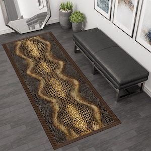 Op de afbeelding: Een rechthoekig tapijt met een slangenprint in bruin- en goudtinten. Het tapijt heeft een donkerbruine rand en ligt op een grijze houten vloer. Een zwarte bank en decoratieve planten zijn op de achtergrond te zien.