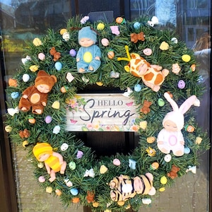 May include: A green spring wreath decorated with plush baby dolls, colorful eggs, and a sign that reads "Hello Spring".