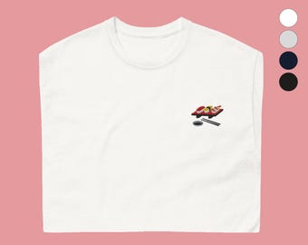 May include: White t-shirt featuring an embroidered sushi design. The design shows sushi pieces in red, yellow, and white on a red tray, with a small black dish and chopsticks. The shirt has a round neck.