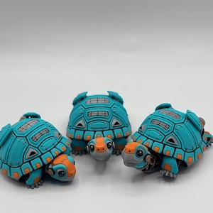 May include: Four teal and orange turtle figurines with a mechanical design. The turtles have detailed shells, with orange accents on their heads, feet, and shell edges. The figurines are arranged on a white surface, with one small turtle in the centre.