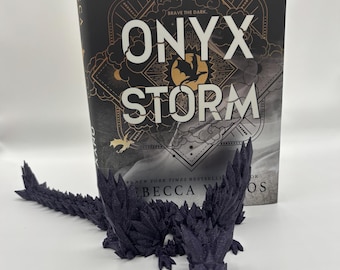May include: A dark purple dragon figurine with a crystalline design is positioned in front of a book titled "Onyx Storm" by Becca Wraos. The book cover is black with gold and white text. The dragon is detailed and 3D printed.