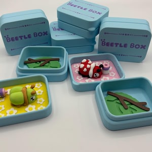 Puede incluir: Contenedores azules claros "Beetle Box" con varias escenas en miniatura en el interior. Una caja presenta una oruga verde con una botella, otra una mariquita roja y otras hojas y ramas.