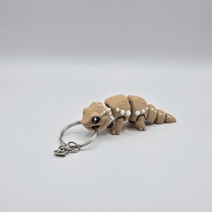 Peut inclure: Un porte-clés en forme de gecko brun et blanc avec un anneau de clé argenté. Le gecko a un corps segmenté et des taches blanches.