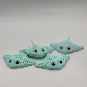 Puede incluir: Cuatro figuras de cerámica de manta raya de color verde azulado claro con alas texturizadas y caras de dibujos animados con ojos azules y detalles blancos. Las mantarrayas están dispuestas sobre una superficie blanca.