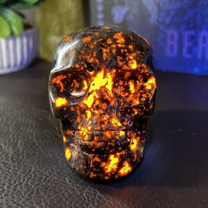 May include: A skull-shaped decorative object with a black base and vibrant orange and yellow patterns. The skull has detailed facial features and a polished surface, creating a striking contrast of colors and textures. This unique piece is ideal for home decor.
