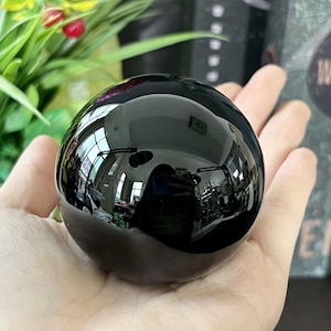 以下が含まれることがあります： 周囲の環境を反射する、研磨された黒曜石の球体。球体の滑らかで光沢のある表面は、鏡のような効果を生み出します。球体は手で持たれており、背景には緑と他のオブジェクトがあります。