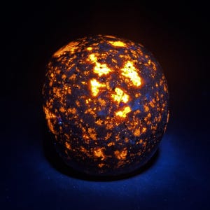 May include: A glowing, spherical sodalite stone. The dark blue stone is speckled with bright orange and yellow patches, creating a celestial appearance. The stone is illuminated against a dark background, highlighting its unique color and texture.