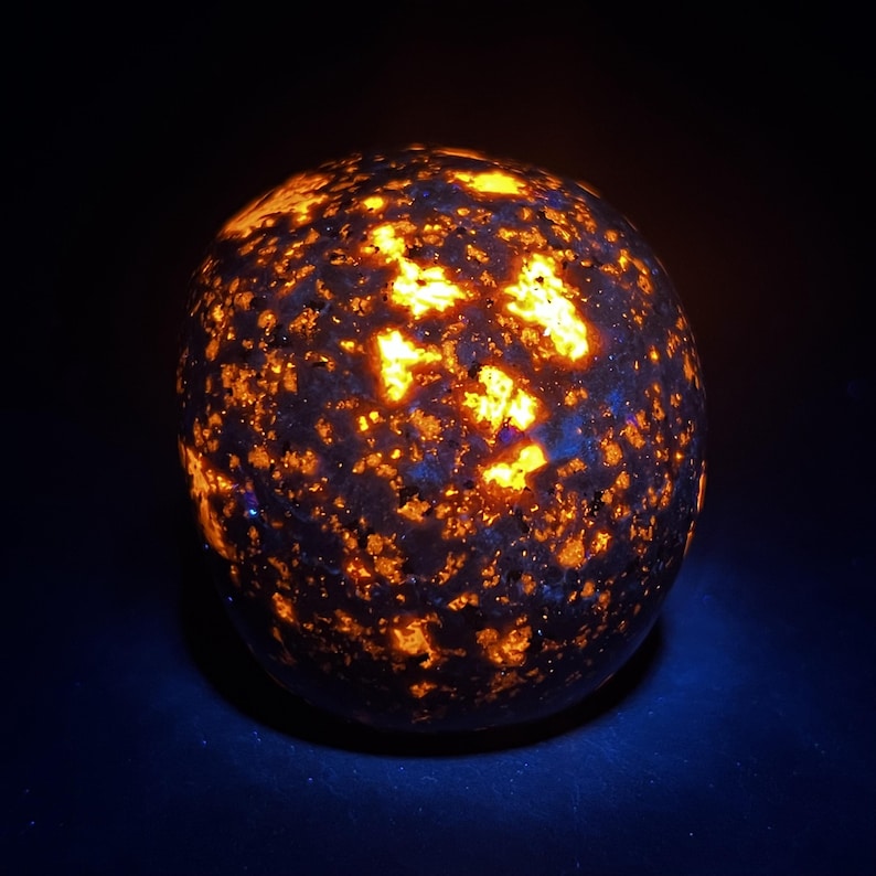 May include: A glowing, spherical sodalite stone. The dark blue stone is speckled with bright orange and yellow patches, creating a celestial appearance. The stone is illuminated against a dark background, highlighting its unique color and texture.