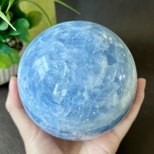May include: A large, spherical, light blue crystal ball. The sphere is held in a hand, and the surface has a cloudy, textured appearance. The crystal ball is a decorative item.