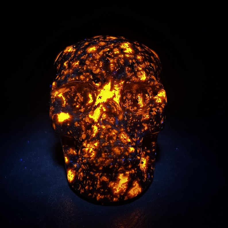 May include: A glowing skull sculpture with a fiery orange and yellow pattern. The skull's surface has a textured appearance, with dark blue and black accents. The background is a deep black, highlighting the skull's vibrant colors.